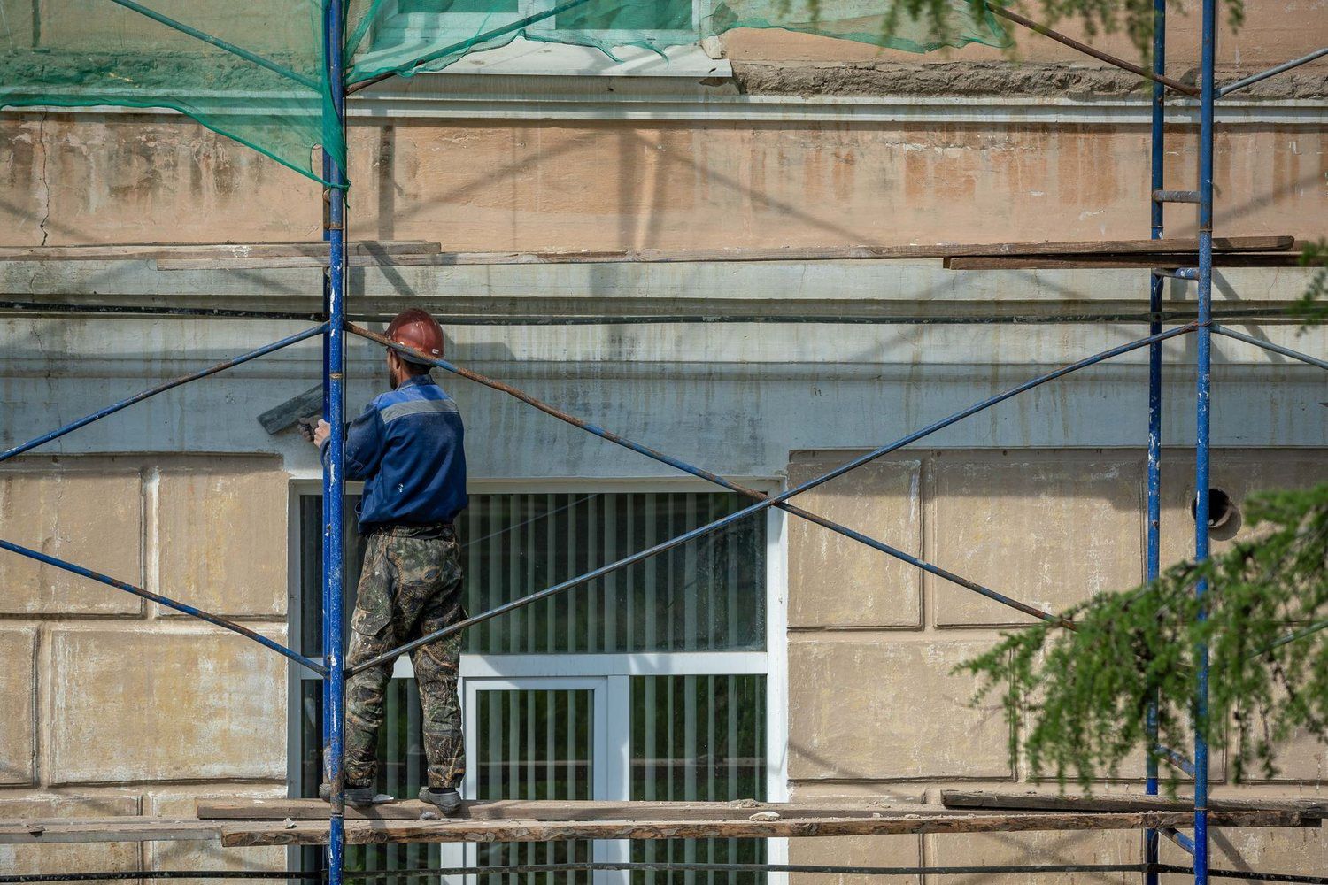 Muncitor cu cască pe o schelă, lângă ferestrele unei fațade în renovare
